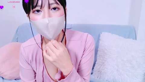 Media: Video of an Asian woman with short black hair, wearing a pink cardigan and a white surgical mask, sitting on a light blue couch with a fluffy white pillow.