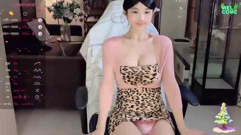 Media: A video of an Asian woman with fair skin, wearing a leopard-print dress and a sheer pink top, sitting on a gaming chair in a modern living room.