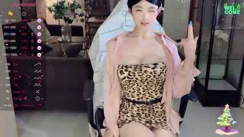 Media: Video of a young Asian woman with fair skin, black hair, wearing a leopard print mini dress and pink jacket, sitting on a gaming chair, making a peace sign. Background shows a modern living room with a TV displaying chat messages.