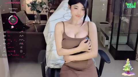 Media: Video of an Asian woman with fair skin, dark hair, and medium breasts in a brown dress, sitting in a chair in a modern living room with a TV showing a game.