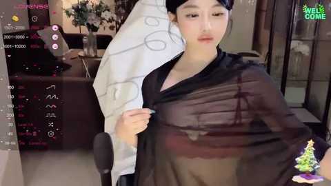 Media: A video of an Asian woman in a black sheer dress, holding a white towel, with a smartphone camera in the foreground, showing a cozy indoor setting with a Christmas tree.