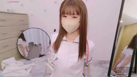 Media: Video of a young East Asian woman with long brown hair in a white nurse's outfit, wearing a face mask, standing in a cluttered, sparsely furnished bedroom with a mirror and dresser.