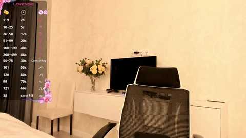 Media: Video of a modern, minimalist bedroom with beige walls, a white desk, black chair, and a flat-screen TV, adorned with a vase of yellow and white flowers, and a dark purple curtain with a digital clock.