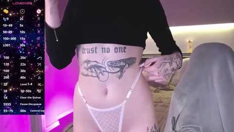 Media: Video of a light-skinned woman with a tattoo of a snake and \"trust no one\" on her abdomen, wearing a black crop top and white lace panties, standing indoors with a purple and pink background.