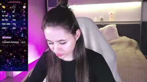 Media: Video of a young woman with long brown hair tied back, wearing a black sweater, sitting on a bed with beige blanket, in a modern bedroom lit by purple and white lights.