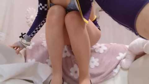 Media: Video of a woman in a purple jacket, sitting on a white bed with pink flower-patterned sheets, wearing blue panties and black high heels.