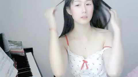 Media: Video of an Asian woman with long black hair, wearing a white polka dot camisole, adjusting her hair in front of a piano.