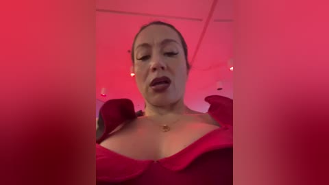 Media: Video of a light-skinned woman with dark hair, wearing a red, off-shoulder dress, with a sultry expression under pink lighting.