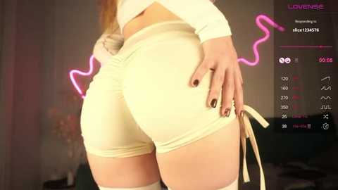 Media: Video of a curvy woman in tight, yellow leggings and a white top, hand on her backside, in a dimly-lit room with a neon pink heart and a digital display in the background.