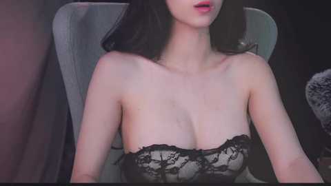 Media: Video of a fair-skinned, slim Asian woman with long black hair, wearing a black lace bra, sitting on a grey chair, indoors.