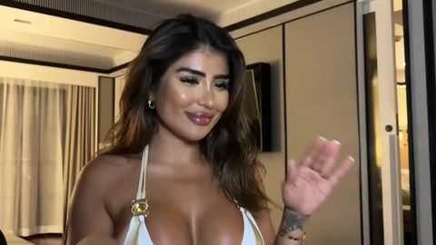 Media: Video of a smiling, voluptuous woman with long, wavy brown hair, wearing a white bikini top, standing in a modern, well-lit room with beige curtains and a TV.