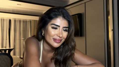 Media: Video of a smiling woman with long, wavy dark hair, medium skin tone, wearing a white top, in a modern hotel room with beige curtains and a flat-screen TV on the wall.