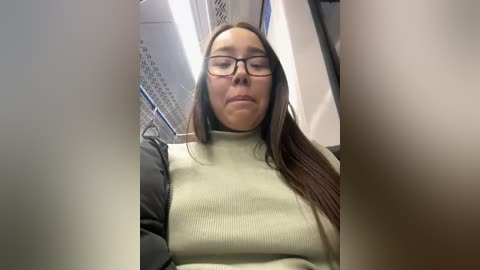 Media: Video of a young woman with long brown hair, wearing glasses and a green sweater, sitting in a subway car. The background features metal railings and a white ceiling.