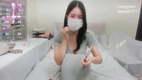 Media: Video of a woman in a hospital bed, wearing a mask, holding a thermometer, with a digital display showing her vital signs, in a white room.