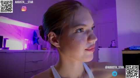 Media: Video of a Caucasian woman with light skin and brown hair tied back, wearing a white tank top, sitting in a dimly lit, modern kitchen with purple LED lighting.