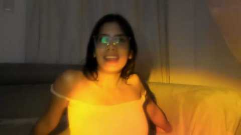 Media: Video of a smiling woman with long black hair, wearing glasses and a yellow top, illuminated by warm yellow light in a dimly-lit room.