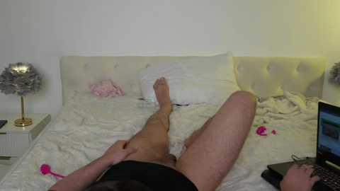 Media: Video of a man lying on a bed, wearing black shorts, holding his penis, surrounded by pink vibrators and a laptop.