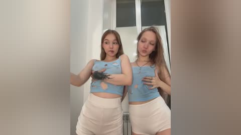 Media: Video of two young, slim women with light skin, long brown hair, wearing blue crop tops and white shorts, standing close in a hallway, making kissy faces.