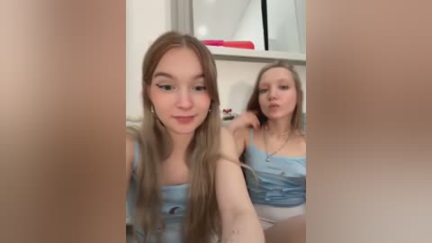 Media: Video of two young women with long blonde hair, wearing light blue tank tops and white shorts, sitting on a bed with a mirror in the background.