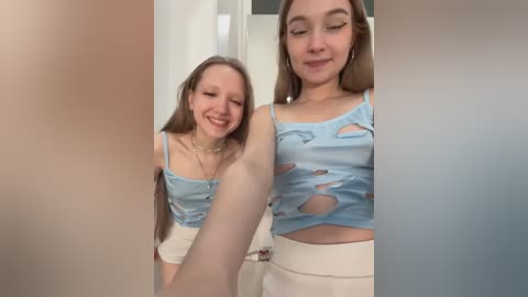 Media: Video of a young woman with light skin and long brown hair, wearing a ripped blue crop top and white shorts, smiling and posing indoors.