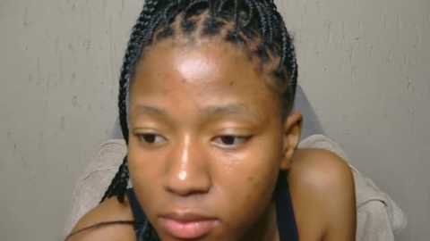 Media: Video of a young Black girl with medium-brown skin, braided hair, and a serious expression, wearing a black top against a plain beige wall.