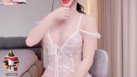 Media: Video of an Asian woman with fair skin, wearing a sheer, white lace lingerie bodysuit, red lipstick, and black hair, sitting on a grey couch.