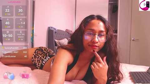 Media: Video of a young woman with long brown hair, wearing glasses, black bra, and leopard print shorts, lying on a bed in a cozy bedroom with a white door, a clock, and a laptop.