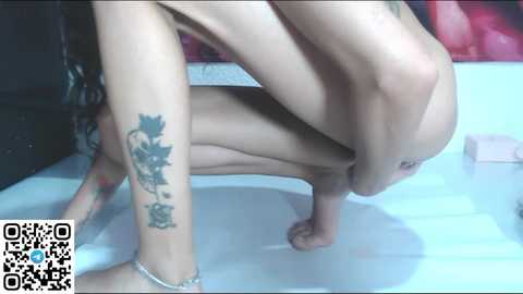 Media: Video of a nude, light-skinned woman squatting, showcasing a floral tattoo on her left calf. Background features a white tiled floor and a pink object.