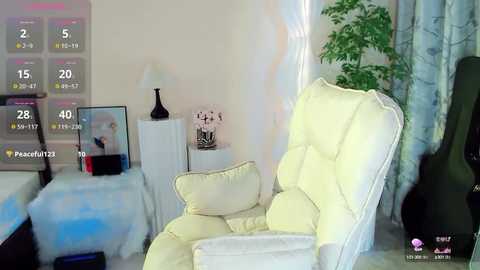 Media: Video of a cozy bedroom featuring a cream armchair, a bed with a blue blanket, a white nightstand with a lamp, and a green plant.