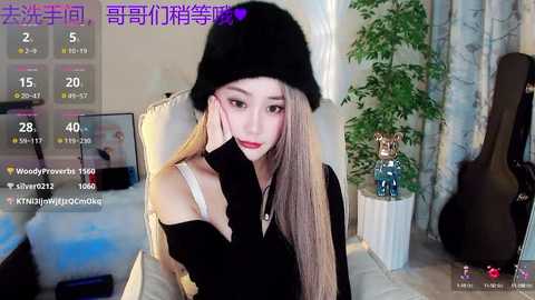 Media: Video of a young East Asian woman with long, straight hair, wearing a black beanie, black sweater, and a white bralette, seated in a cozy room with a potted plant and musical instruments.