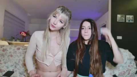 Media: Video of two women in a bedroom: one blonde with a pink top, the other brunette with long hair, both smiling.