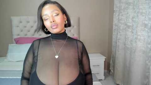 Media: Video of a Black woman with a medium skin tone and short black hair, wearing a sheer black top revealing her large breasts, in a bedroom with a white tufted headboard, white nightstand, and silver curtains.