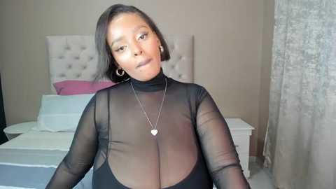 Media: Video of a curvy Black woman with medium skin tone, wearing a sheer black top that reveals her large breasts, standing in a bedroom with a white bed and beige wall.