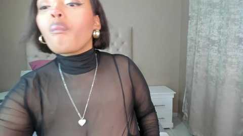 Media: Video of a dark-skinned woman with a medium build, wearing a sheer black top revealing her bra and a heart pendant necklace, making a pouty face in a beige bedroom with white furniture.