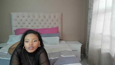 Media: Video of a young Black woman with long hair, wearing a black sheer top, standing beside a neatly made bed with pink and white pillows, in a softly lit, modern bedroom.