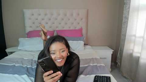 Media: Video of a smiling Black woman with medium brown skin, lying on a bed in a modern bedroom, wearing a black sheer top, holding a smartphone, surrounded by pink and white bedding.