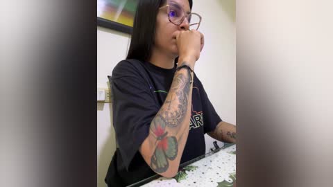 Media: Video of a woman with long black hair, glasses, and tattoos on her arm, wearing a black t-shirt, sitting at a table in a dimly lit room.