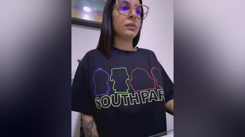 Media: Video of a young woman with long dark hair, wearing glasses and a black T-shirt with colorful silhouettes of heads and \"SouthPark\" text.