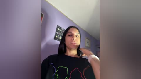 Media: Video of a young woman with straight black hair, wearing a black graphic t-shirt, standing in a room with light purple walls and a framed picture.