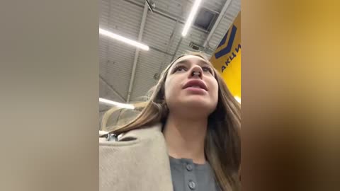 Media: Video of a young woman with long brown hair, wearing a beige coat over a blue shirt, standing in a dimly lit, industrial setting with exposed metal beams and fluorescent lights.