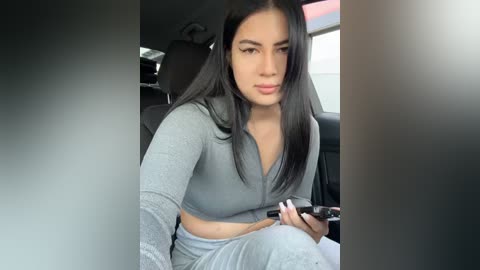 Media: Video of a young woman with long, straight black hair, wearing a grey crop top and matching pants, sitting in a car, holding a smartphone, with a neutral expression.