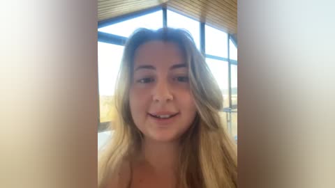 Media: Video of a young Caucasian woman with long, wavy blonde hair, light skin, and a slight smile, standing indoors near a large window with wooden paneling.