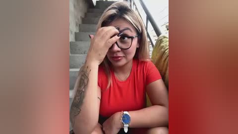 Media: Video of a young Asian woman with straight, shoulder-length blonde hair, wearing black-framed glasses and a red t-shirt, sitting on a staircase, adjusting her glasses, with a tattoo on her left forearm.