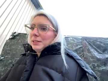 Media: Video of a woman with blonde hair, wearing glasses, a black puffy jacket, and a white hat, standing in front of a grey brick wall and a tarp-covered structure.