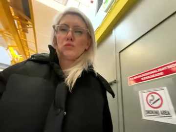 Media: Video of a blonde woman in glasses, wearing a black puffy jacket, standing in a modern, brightly lit underground station.