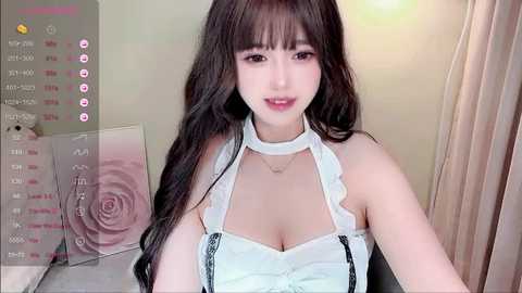 Media: Video of a young Asian woman with long, wavy black hair, wearing a white halter top, smiling. Background features a beige wall with a pink rose print calendar.