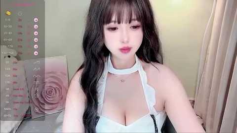 Media: A video of a young East Asian woman with long, dark hair, wearing a white, lace-trimmed halter top, revealing ample cleavage. She's indoors, near a wall with a pink rose design and a calendar.