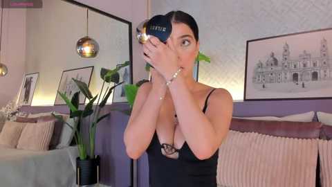 Media: Video of a woman in a black lace bra, holding a camera, in a modern bedroom with framed cityscape art and a green plant.