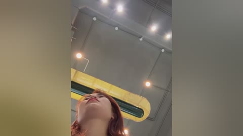 Media: Video of a woman with light skin and long red hair, looking up at a modern, industrial ceiling with bright lights and yellow ventilation ducts.