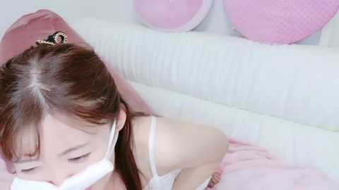 Media: Video of an Asian woman with long brown hair, wearing a white mask, lying on a pink bed with white pillows in a soft, pastel-toned room.